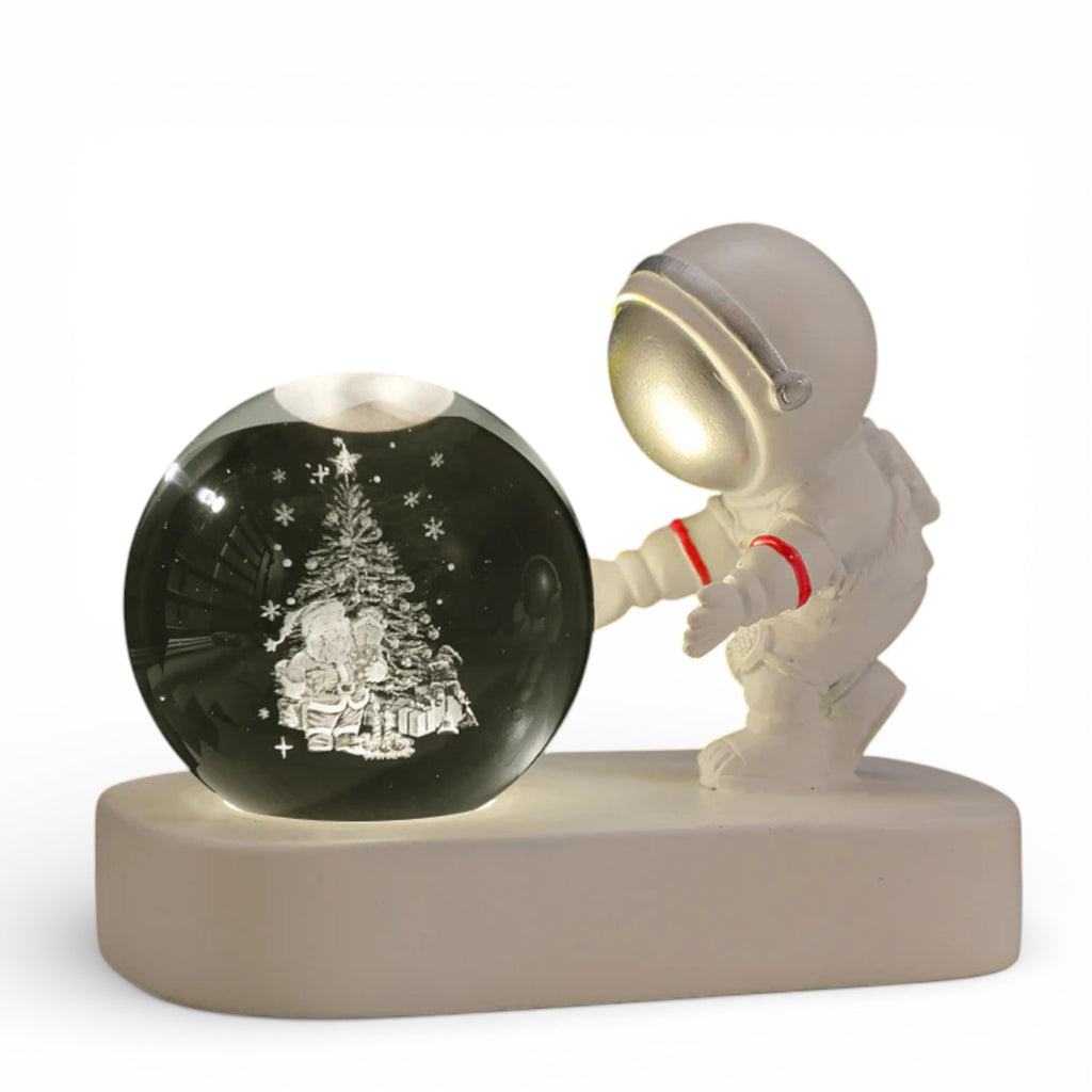 Cute astronaut figurine with a glowing helmet beside a crystal ball engraved with a Christmas tree and gifts